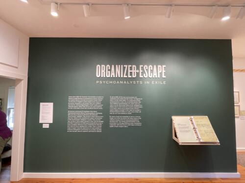 Museum title graphics read "Organized Escape". Custom fabrication of display mounted to wall holding binder of exhibition materials. Interactive display