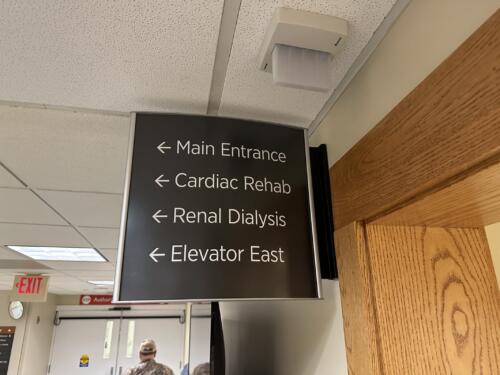Custom fabricated projecting sign for hospital hallway. Sign reads "main entrance, cardiac rehab, renal dialysis and elevator east" with directional arrows.