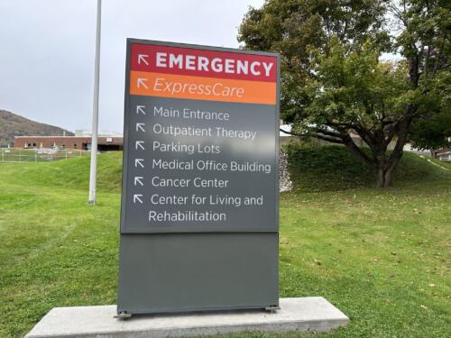 Freestanding road sign for hospital wayfinding. Sign reads "emergency, express care, main entrance, outpatient therapy and parking lots" with directional arrows.