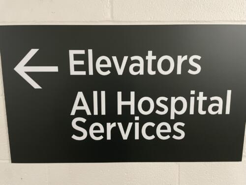 Interior directional sign with left arrow for hospital wayfinding. Sign reads "Elevators and All Hospital Services".